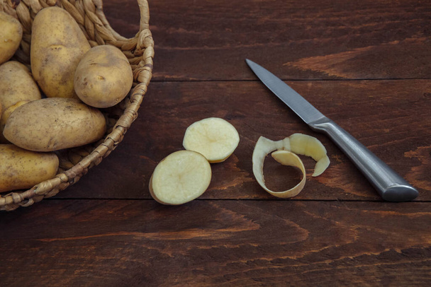 New potatoes in a wicker basket cut in half and peeled on a brown wooden background with a metal knife top view, cooking concept, Ingredients, recipes, lunch and dinner, French fries, soup and chips - Foto, Imagen