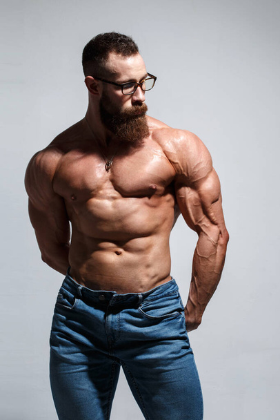 Muscular young man with a beard and glasses with a naked torso isolated on a gray background. - Fotoğraf, Görsel