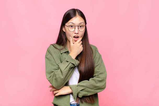 asian young woman with mouth and eyes wide open and hand on chin, feeling unpleasantly shocked, saying what or wow - 写真・画像