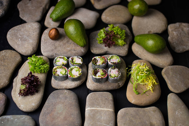 Widok z boku japońskiego sushi rolki bez ryżu z mięsem kraba śniegu (paluszki krabowe) zawinięte w daikon rzodkiewki serwowane na kamieniach morskich. składniki na tle. Całe awokado i mieszanka sałatek bez ryżu.  - Zdjęcie, obraz