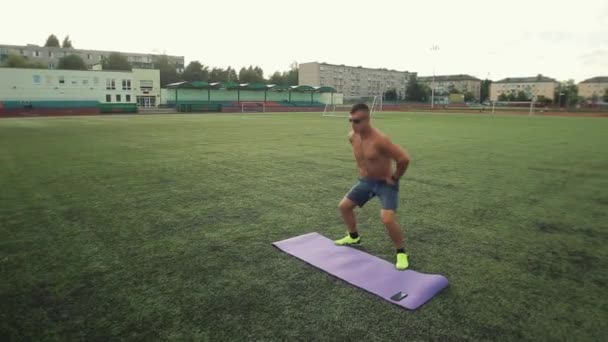 A bodybuilder in sports shorts and sunglasses doing exercises on the football field of the city stadium with a timer on his mobile phone.. Front view. The camera moves behind the athlete - Footage, Video