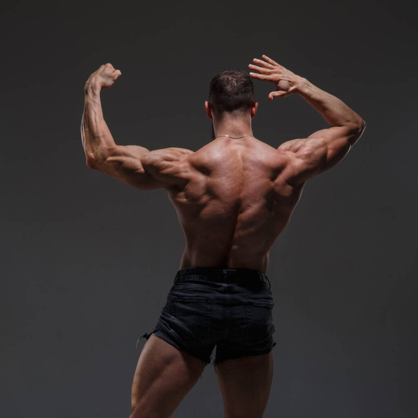 Portrait of a  athletic man with a naked torso showing muscles isolated on a dark gray background, view from the back. - Foto, Imagen