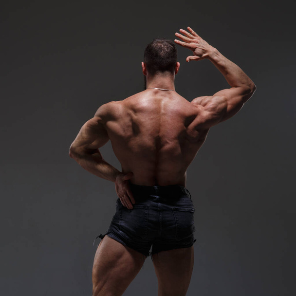Portrait of a  athletic man with a naked torso showing muscles isolated on a dark gray background, view from the back. - Fotografie, Obrázek