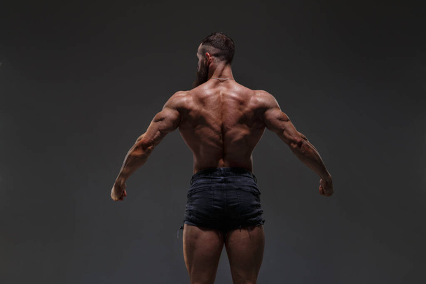 Portrait of a  athletic man with a naked torso showing muscles isolated on a dark gray background, view from the back. - Photo, Image