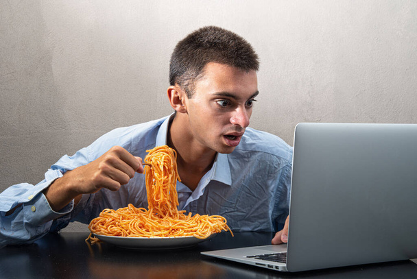 un uomo che mangia spaghetti con salsa di pomodoro e guarda il computer - Foto, immagini