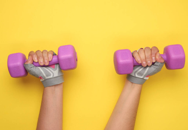 female hand in a pink sports glove holds a purple one kilogram dumbbell on a yellow background - Photo, Image