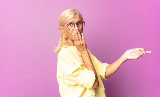 middle age woman feeling happy, shocked and surprised, covering mouth with hand and pointing to lateral copy space - 写真・画像