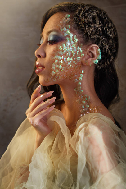 Beauty portrait of a beautiful Asian girl with creative make-up of a mermaid and scales on the face view in profile. - 写真・画像