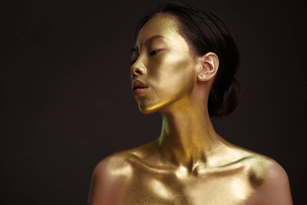 Beauty portrait of a beautiful asian girl with gold paint on her face against a dark background. - Foto, Imagen