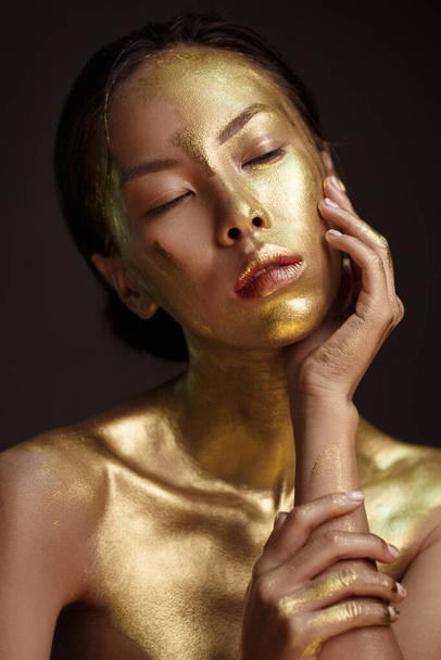 Portrait de beauté d'une belle fille asiatique avec de la peinture d'or sur son visage sur un fond sombre. - Photo, image