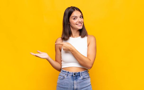young pretty woman smiling cheerfully and pointing to copy space on palm on the side, showing or advertising an object - Foto, Imagen