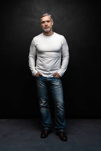 Full body portrait of middle-aged good looking man in white t-shirt posing in front of a black background with copy space. - Fotografie, Obrázek