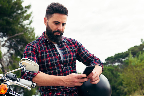 handsome bearded man consulting his mobile phone sitting on his motorcycle with his helmet under his arm, concept of freedom and biker lifestyle, copy space for text - Foto, Imagen