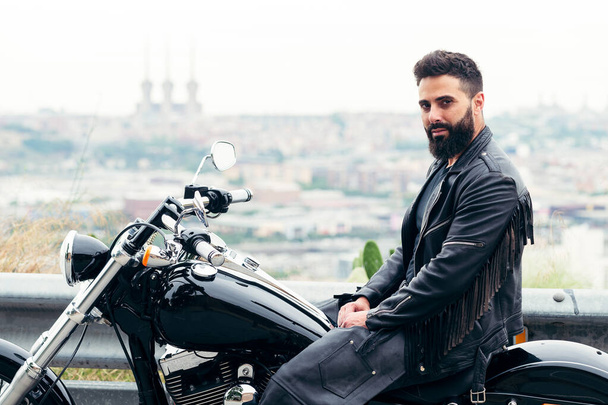 bearded man dressed in leather sitting on his motorcycle with panoramic views of the city, concept of freedom and biker lifestyle, copy space for text - Foto, afbeelding