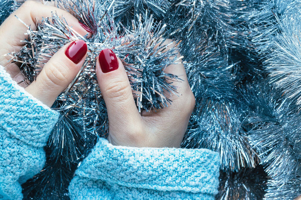 Kobiece dłonie w niebieskim swetrze z dzianiny z pięknym błyszczącym manicure - bordowy, ciemnoczerwony, wiśniowe paznokcie na tle srebrnej girlandy świątecznej - Zdjęcie, obraz