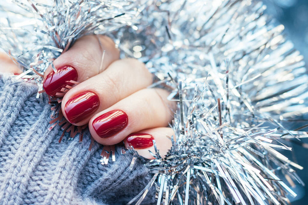 Female hands in a gray knitted sweater with a beautiful glossy manicure - burgundy, dark red, cherry color nails on background of silver Christmas tinsel garland with copy space. Selective focus - Фото, изображение