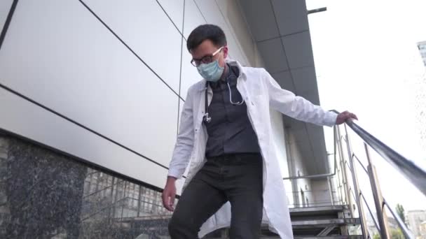 Kaukasischer junger Arzt setzt sich auf die Treppe in der Nähe des Klinikgebäudes, müde und unglücklich reibt er sich Nase und Augen, fühlt sich müde und hat Kopfschmerzen. Konzept für Stress und Frustration im Gesundheitswesen - Filmmaterial, Video