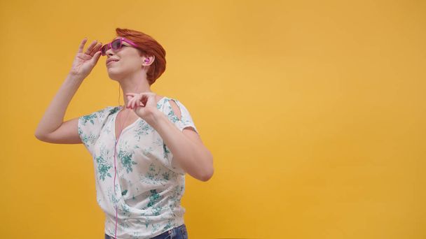 Drôle de femme aux cheveux rouges avec des lunettes de soleil roses dansant sur le fond jaune. Femme dans sa quarantaine - Photo, image
