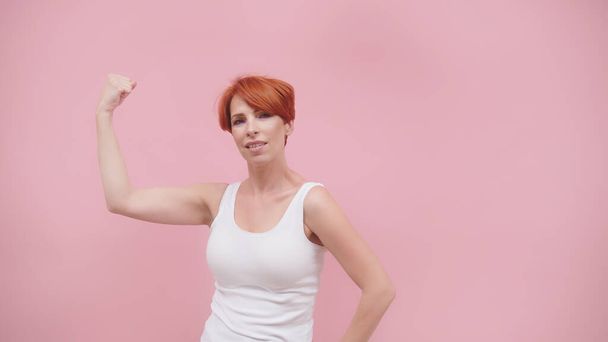 Красивая возбужденная рыжая женщина средних лет разминает свои мышцы, изолированные на розовом фиолетовом фоне - Фото, изображение