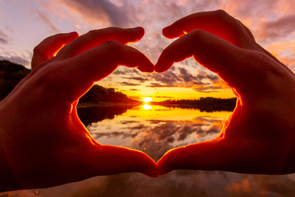 Αρσενικά χέρια κάνουν μια χειρονομία σε σχήμα καρδιάς με φόντο το ηλιοβασίλεμα ή την αυγή σε ένα ποτάμι ή λίμνη το καλοκαίρι. - Φωτογραφία, εικόνα