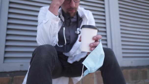 Ręce z bliska z filiżanką kawy i ochronną maską medyczną relaksującą po powrocie pacjentów do zdrowia i ciężkiej pracy. Pracownik służby zdrowia czuje się zmęczony po ochronie i walce covid 19 gorącym, energetyzującym napojem - Materiał filmowy, wideo
