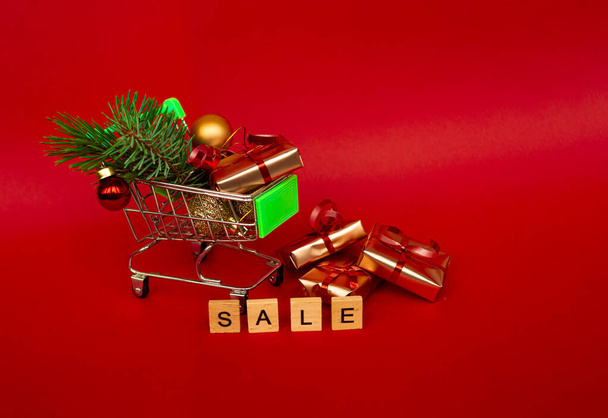 Grocery basket with golden gift boxes and Christmas balls, a sprig of Christmas tree and English letters SALE on a red background banner with copy space. Xmas sale. Christmas shopping online. Winter holiday sales and discounts, seasonal sales, Black  - Фото, зображення