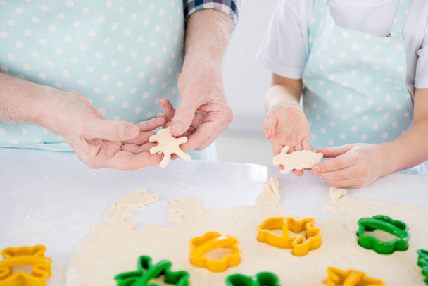 Cropped close-up view of nice hands granddad grandchild preparing cooking cookies sweet snack using cutter animal shape form figure help assistance in modern light white interior kitchen house - Foto, afbeelding