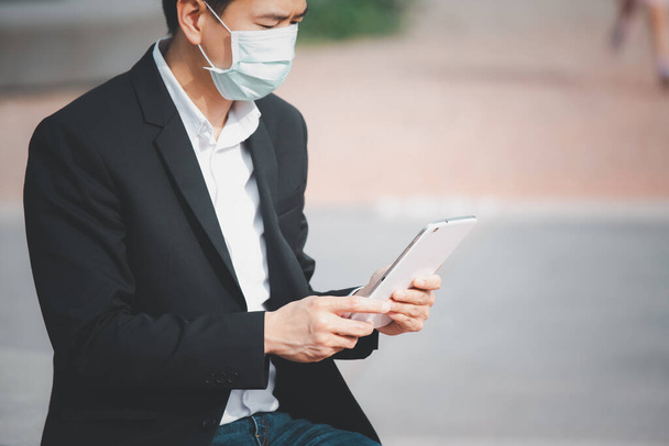 A imagem rosto de um homem de negócios asiático vestindo uma máscara médica para evitar germes, fumos tóxicos, e COVID 19, Ele está lendo e ouvindo as notícias em tablets e jornais, não funcionam no parque. - Foto, Imagem