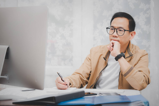 Asian man in casual clothing using a laptop communicates on the internet with the customer at  kitchen table at home. concept work from home. - Photo, Image