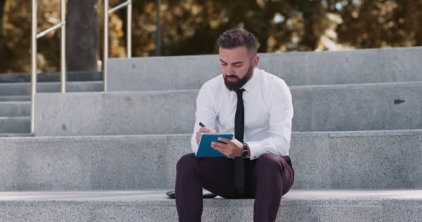 Concentrated businessman creating marketing plan, sitting on stairs in park - Footage, Video
