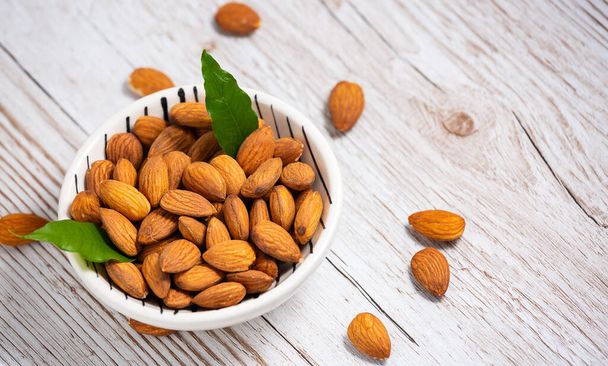 Valkoisessa kupissa suolaan sekoitetut mantelinpähkinät ovat proteiiniruokaa ja terveellisiä elintarvikkeita painonpudotukseen ruokavalioon puun jyvien taustalla.. - Valokuva, kuva