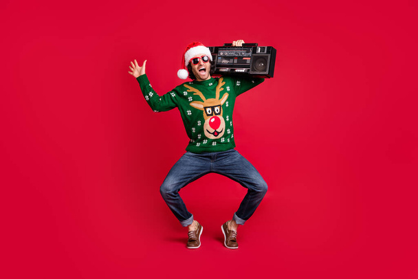 Full length body size view of attractive glad cheerful childish guy wearing Santa hat carrying boombox having fun dancing fooling free time isolated bright vivid shine vibrant red color background - Foto, imagen