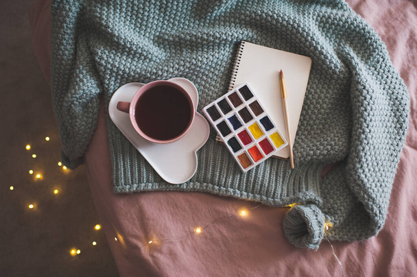 Kupa friss kávé tányéron palettája festékek, vázlatfüzet és kötött pulóver felett izzó karácsonyi fények a háttérben az ágy közelében. Téli szezon. Kreatív koncepció. Felülnézet.  - Fotó, kép