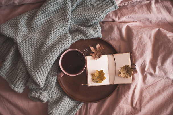 Taza de té negro con cuaderno abierto en bandeja de madera y suéter de punto en la cama de cerca. Temporada de otoño. Buenos días. Hora del desayuno.  - Foto, Imagen