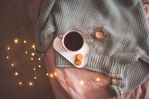 Mug of fresh black tea or coffee with tasty cookies with candle and glowing lights on knitted wool sweater in bed close up. Good morning. Breakfast time. Autumn season. Top view.  - Photo, Image