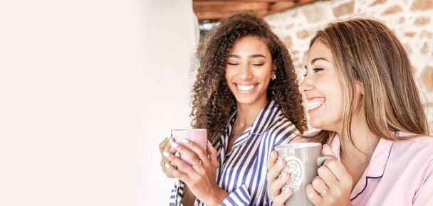 Мульти-этнические лучшие друзья женщины в пижаме улыбаются держа чашку чая с белым копировальным пространством слева - Концепция: оставайтесь дома и наслаждайтесь жизнью - Фото, изображение
