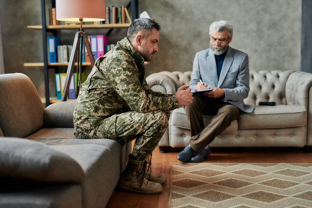 Healing process. Full length shot of middle aged military man looking away during therapy session with psychologist. Soldier suffering from depression, psychological trauma. PTSD concept - Photo, Image