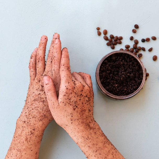Top view of female hands and a jar with roasted coffee bean scrub and sea salt on mint background in square format. Rejuvenating scrub in round jar for skin exfoliation. - Photo, Image