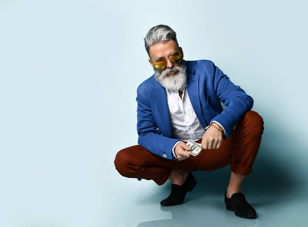 Elderly man in white shirt, jacket, brown pants, black loafers and sunglasses. Squatting, showing cash, posing on blue background - Фото, изображение