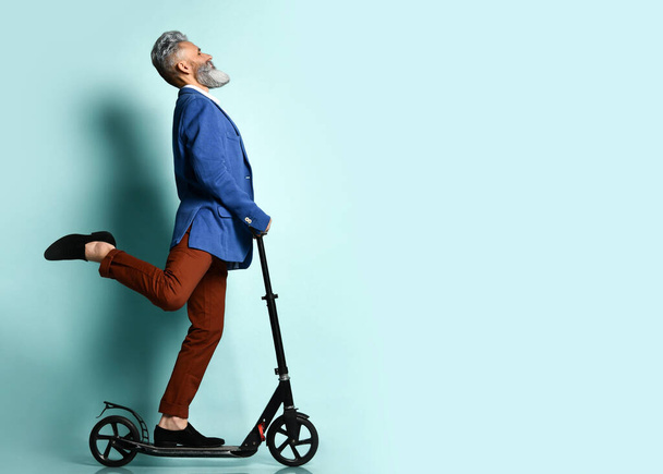 Gray-haired male in white shirt, jacket, brown pants and loafers. Riding black scooter and posing sideways on blue background - Fotó, kép