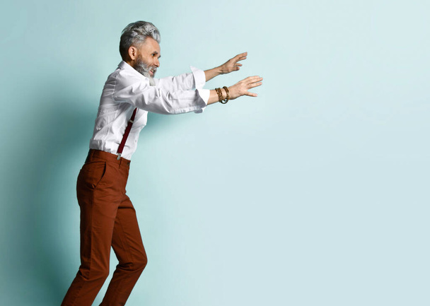 Aged man in white shirt, brown pants and suspenders, black loafers. He runs looking back, raised his hand up, blue background - Fotografie, Obrázek