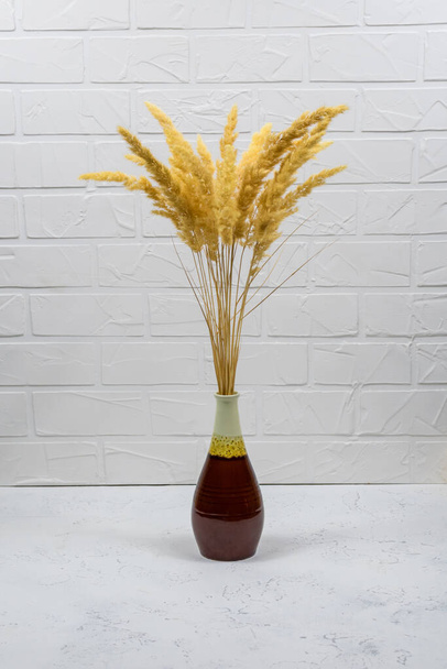 A bouquet of dry spikelets in a brown ceramic vase on a wooden table against a white brick background. Stylish home decor. Modern interior design - Foto, imagen