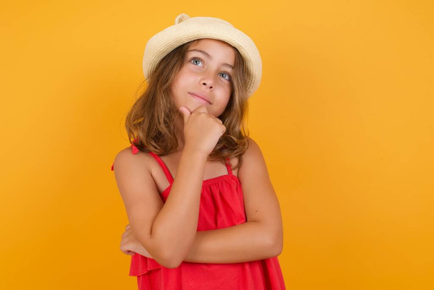 schattig klein meisje dragen rode jurk denken en poseren op gele achtergrond  - Foto, afbeelding