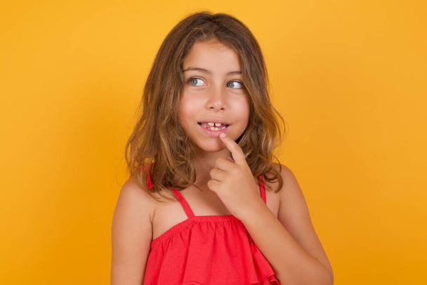 schattig klein meisje dragen rode jurk denken op gele achtergrond  - Foto, afbeelding