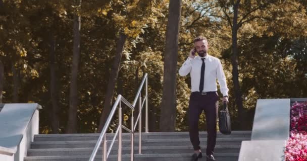 Hombre de negocios positivo hablando por teléfono y caminando en el parque de otoño, sosteniendo la chaqueta en las manos, disfrutando del clima cálido - Imágenes, Vídeo