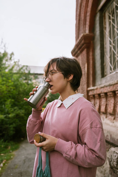 Junge erwachsene Frau mit kurzen Haaren trinkt aus Metall nachhaltiges Wasser im Freien - Foto, Bild
