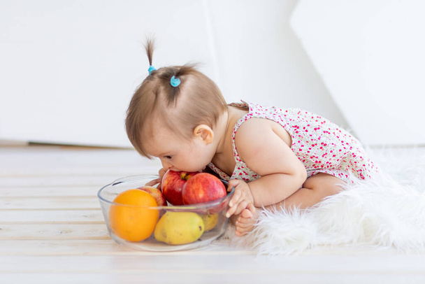 Küçük bir kız bir tabak meyveyle parlak bir odada oturuyor ve bir elma yiyor. - Fotoğraf, Görsel