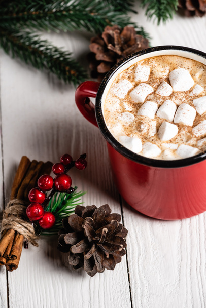 Cup of cocoa with marshmallows near cinnamon sticks with red beads and pine cone - Photo, Image