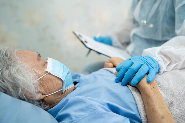 Dokter draagt PBM pak nieuw normaal te controleren Aziatische senior of oudere oude dame patiënt met een gezichtsmasker in het ziekenhuis voor infectie te beschermen Covid-19 Coronavirus. - Foto, afbeelding