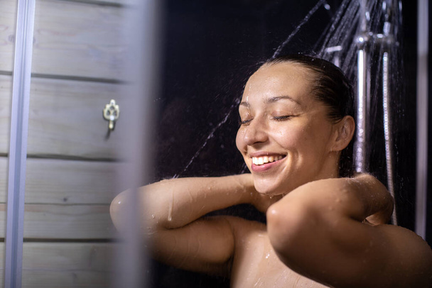 sorrindo mulher nua desfrutando de água corrente de pé em casa de banho fechar os olhos - Foto, Imagem